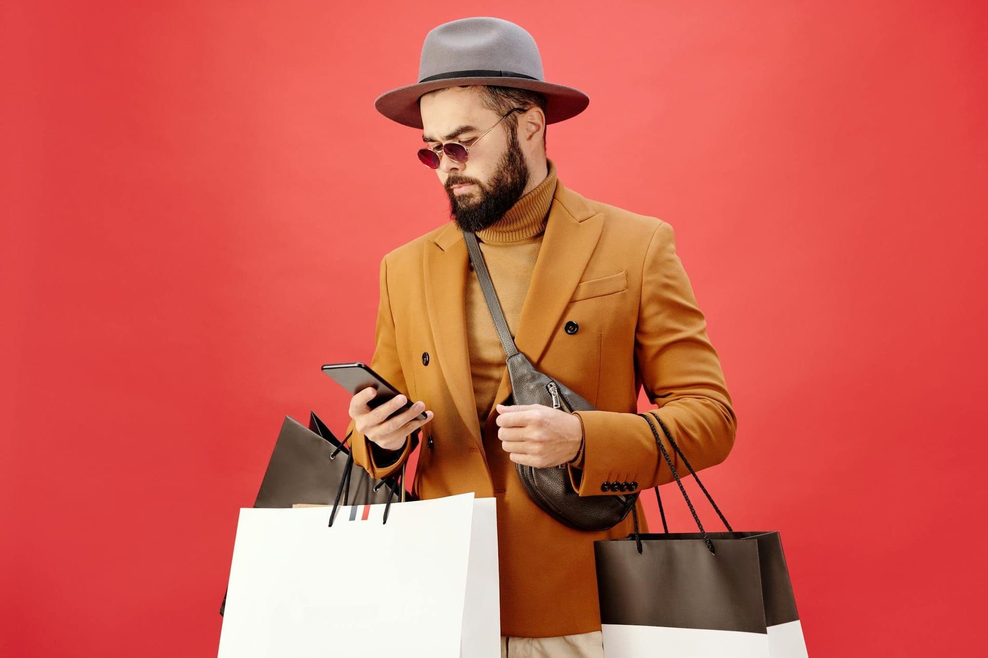 Bearded shopper in a mustard coat checking his phone while carrying shopping bags against a red backdrop