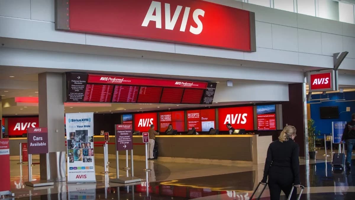 Traveler wheeling luggage toward a staffed Avis rental counter in an airport terminal