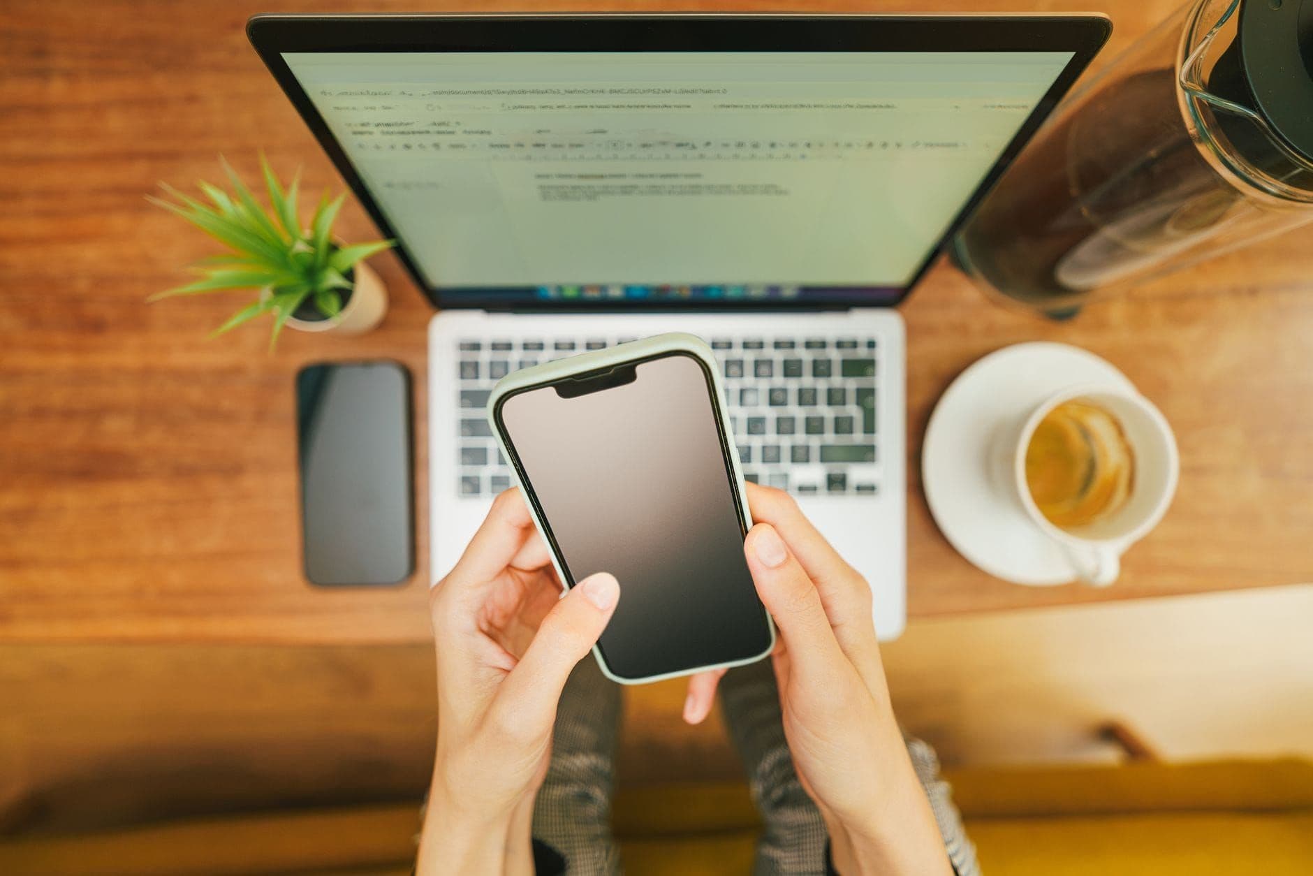 Hands holding a smartphone over a laptop beside a cup of coffee