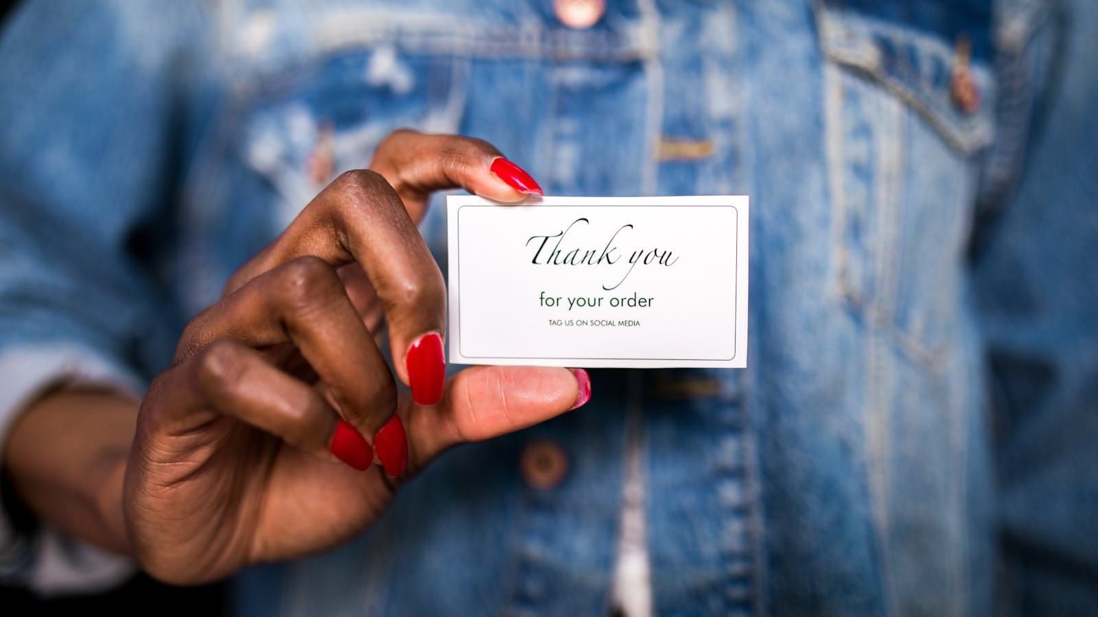 Hand with red nails holding a Thank you for your order card in front of a denim jacket