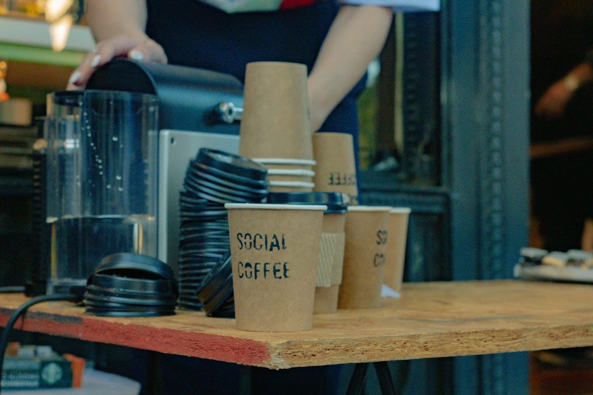 Kraft takeaway cups stencilled Social Coffee stacked on a cafe counter beside an espresso machine