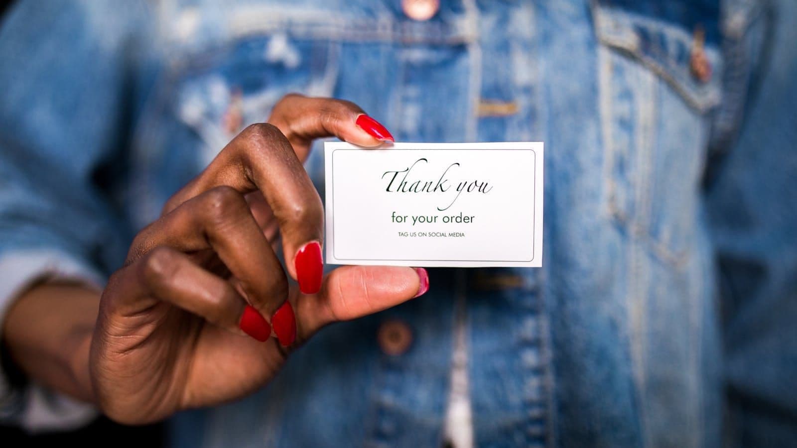 Hand with red nails holding a Thank you for your order card in front of a denim jacket