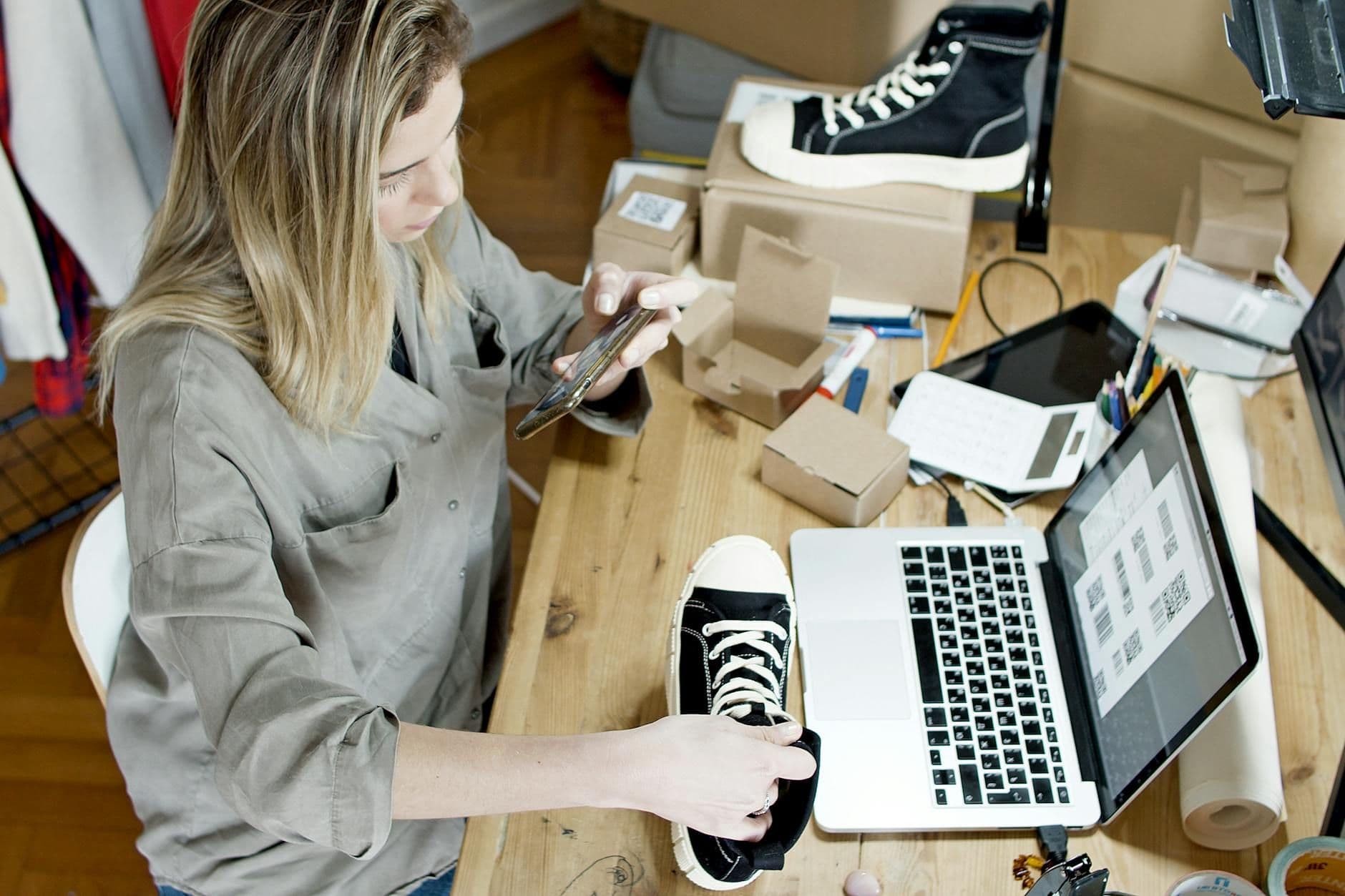 Small-business owner photographing a black sneaker with her phone at a desk piled with shipping boxes